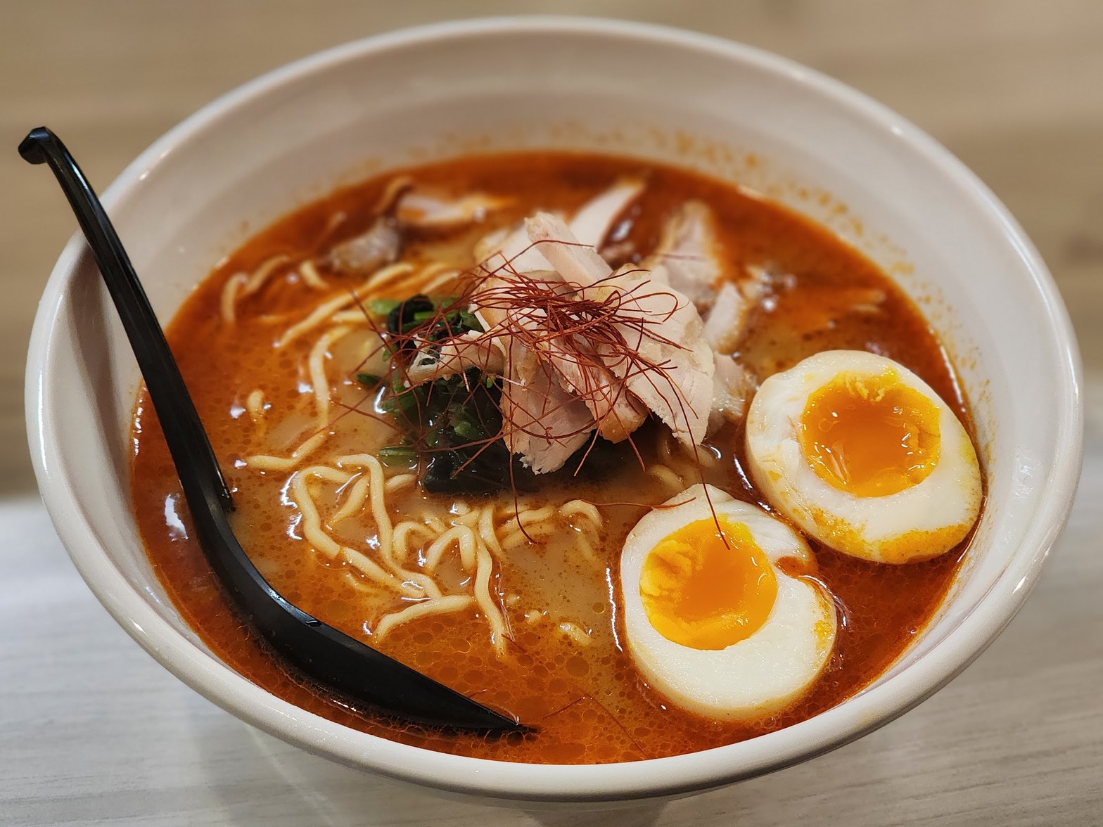 赤みがかったスパイシー醤油スープに麺・チキン・半熟卵2個・糸唐辛子をのせた白い丼と黒いレンゲ